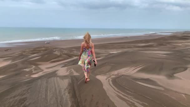 Blonde woman walking along the top of a dune and stops to take a picture with her smartphone. — стоковое видео