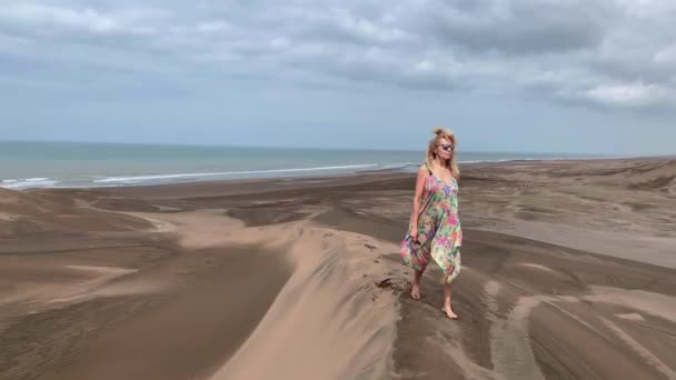 Blonde woman walking on a dune with sunglasses and mobile phone in her hand. — Stockvideo