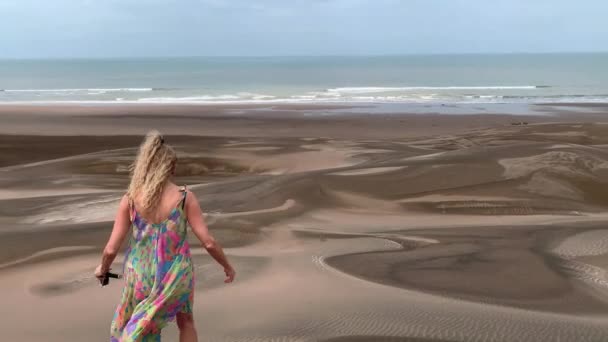 Vista de trás de uma mulher loira em um vestido de praia colorido andando por uma duna em direção à praia. — Vídeo de Stock