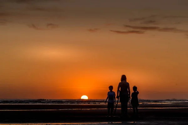 两个孩子抱着拐杖在海滩观看落日的女子背光 — 图库照片