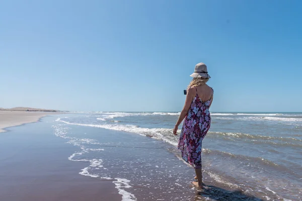 Woman in summer clothes strolling barefoot along a windy beach recording with a mobile-