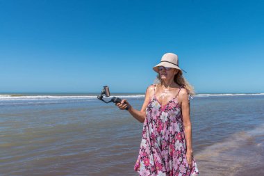 Sahilde duran şapkalı bir kadın cep telefonuyla video çekiyor.