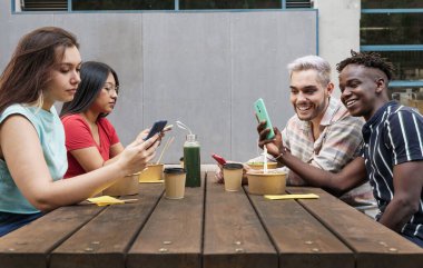 Akıllı telefon kullanan, komik fotoğraf ya da video içeriğini sosyal ağlarda paylaşan mutlu genç melez arkadaş grubu. İşten sonra açık büfe bar masasında oturup yemek yemek.
