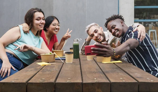 Glade, unge venner som bruker smarttelefon for å ta selfiefotografier som del i sosiale medier. Sitte utendørs etter et barbord i byen med mat – stockfoto