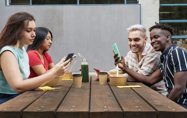 Glade, unge venner som bruker smarttelefon og deler morsomme bilder eller videoinnhold i sosiale nettverk. Sitte på et utendørs barbord i byen etter jobb med å ta bort mat – stockfoto