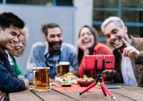 Ryhmä onnellisia monikansallisia ystäviä, jotka nauttivat online-videopuhelun tekemisestä matkapuhelimella oluen baarissa syntymäpäivän kunniaksi - Keskity matkapuhelimeen — kuvapankkivalokuva