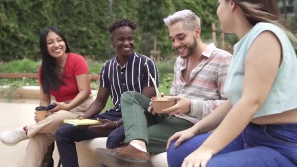 Grupa młodych kosmopolitycznych przyjaciół wielokulturowych studentów lub kolegów cieszących się rozmową podczas jedzenia jedzenia jedzenia na wynos ulicznego jedzenia w parku miejskim. Skup się na Afrykaninie. — Wideo stockowe