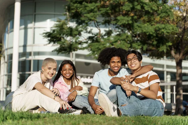 Ryhmä muotokuva nuorten monipuolinen yliopisto opiskelija ystäviä istuu ruohoa kampuksella puistossa — kuvapankkivalokuva