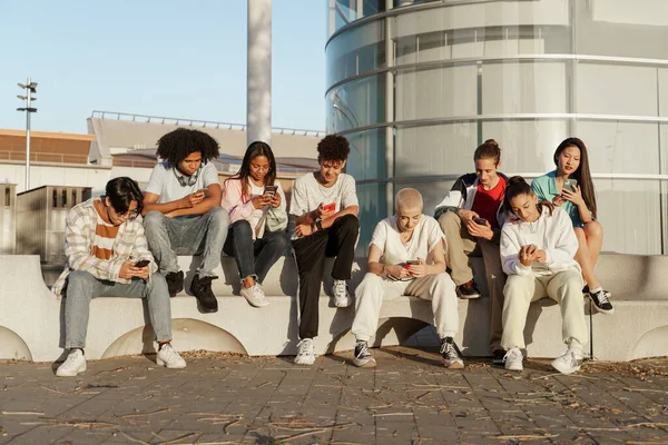 Monikulttuurinen ryhmä nuoria trendikkäitä ystäviä matkapuhelimia ja sosiaalista mediaa käyttäen - Opiskelijat istuvat modernilla kaupunkialueella ja kirjoittavat älypuhelimilla — kuvapankkivalokuva