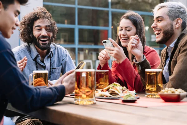 En gruppe lykkelige venner som nyter en øl og mat på utendørs bar pub etter jobb, og som deler gode øyeblikk i sosiale medier med smarttelefoner – stockfoto