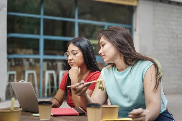 Nuori keskittynyt latino nainen työskentelee kannettavan kun taas naispuolinen kollega syö terveellistä ottaa aasialainen nuudelit ulkona pöydän kaupungissa — kuvapankkivalokuva