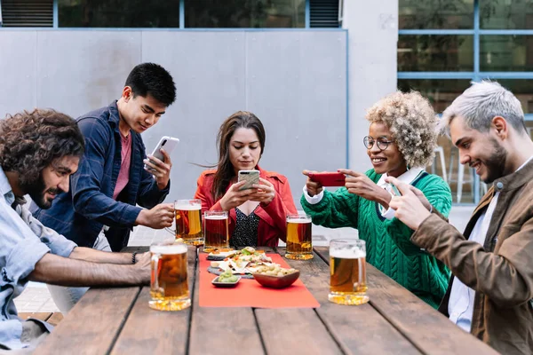Ryhmä onnellisia monikansallisia ystäviä älypuhelimilla, jotka ottavat kuvan ruoasta ja juomasta ulkona baarissa tai pubissa jakaakseen sen sosiaalisen median sovelluksissa — kuvapankkivalokuva