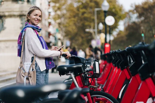 Vrouw neemt een rode fiets in een fietsverhuur station in de stad met fiets delen app. Ecologische manier van woonwerkverkeer in de stad — Stockfoto