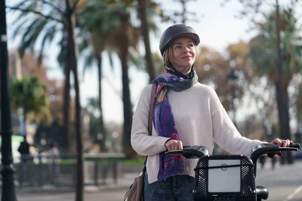 Jonge zelfverzekerde vrouw met fietshelm duwen elektrische huur delen fiets in een stadspark met palmbomen. Eco en gezonde forensen — Stockfoto