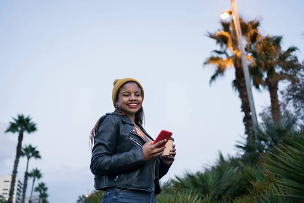Mote ung afroamerikansk kvinne i stilig skinnjakke med smarttelefon som nyter nattlys i byparken – stockfoto
