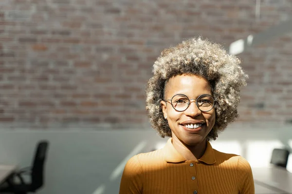 Portrett av stilige, glade og selvsikre afroamerikanske unge profesjonelle kvinner med blond afrofrisyre, briller og gule klær på et moderne kontor med mursteinsvegger – stockfoto