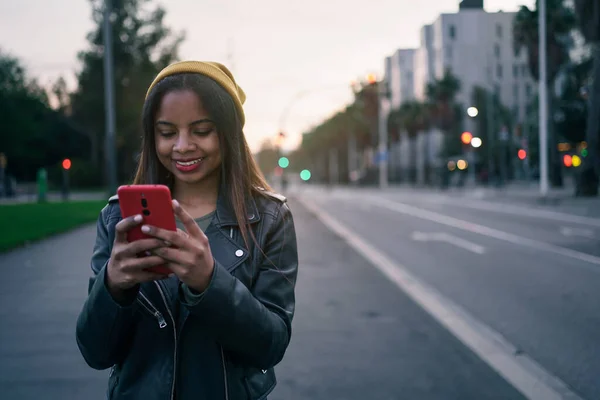 Mote ung afroamerikansk kvinne i stilig skinnjakke med felles bilder på sosiale medier med smarttelefon i byen ved solnedgang – stockfoto