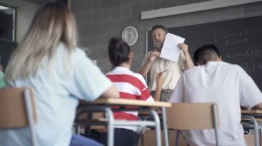 Friendly Professor explaining maths exam exercise in high school classroom and teenager student raising hand to ask question