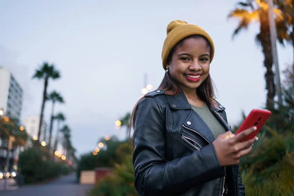 Smiling African American female holding smartphone standing on city street at sunset – stockfoto