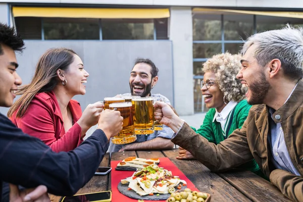 Onnellisia nuoria ystäviä paahtaminen tuoppi olutta juhlii ystävyyttä yhdessä ulkona baarissa — kuvapankkivalokuva