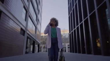 CBD bölgesindeki ofis binaları arasında yürüyen kendine güvenen siyahi iş kadını iş arkadaşlarıyla telefonda konuşuyor.