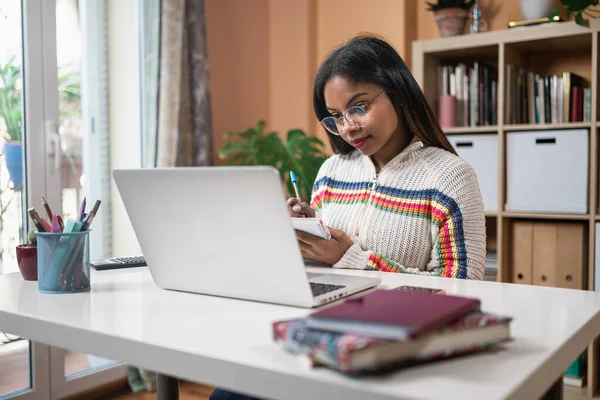 Afrikkalainen amerikkalainen naispuolinen opiskelija istuu pöydässä osallistumalla online-luokkaan kotona — kuvapankkivalokuva