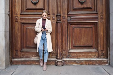 Akıllı telefon ekranına bakan çekici zarif bir kadın ahşap kapının üzerinde duran internetten mesaj atıyor.