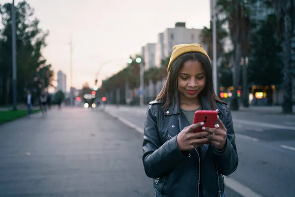 Huoleton nuori musta nainen tyylikkäässä nahkatakissa älypuhelimella kaupungissa illalla — kuvapankkivalokuva