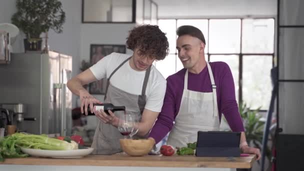 Feliz casal gay se divertindo em casa, bebendo e cozinhando juntos na cozinha. Homem derramando vinho para seu namorado — Vídeo de Stock
