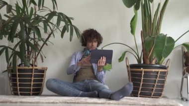 Rahatlamış, kıvırcık saçlı, tablet bilgisayarında internette gezinen genç bir adam, ekranı okurken mutlu bir şekilde yerde oturuyor.
