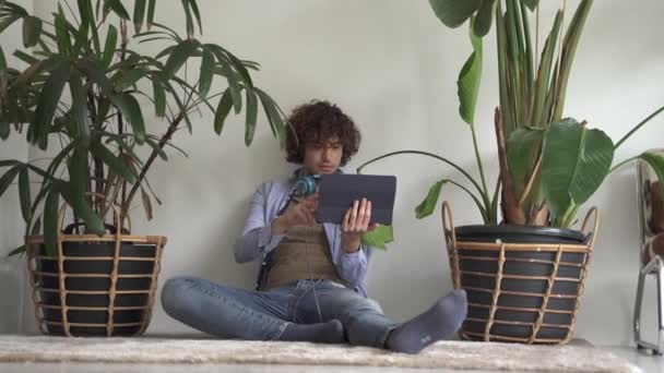 Relaxed young man with curly hair browsing the internet on his tablet computer as he sits on on the floor at home happy as he reads the screen — Stock Video