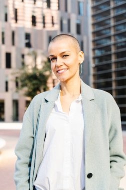 Tıraşlı saçlı şık bir iş kadını kameraya kendinden emin bir ifadeyle bakar.