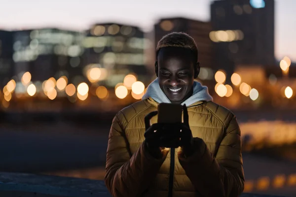Nuori onnellinen afrikkalainen mies käyttää matkapuhelinta kaupungissa yöllä — kuvapankkivalokuva