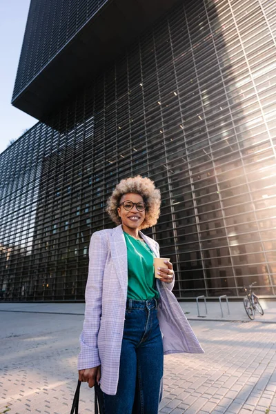 Smilende afroamerikansk forretningskvinne med pose og kaffe stående i byen på solskinnsdag – stockfoto