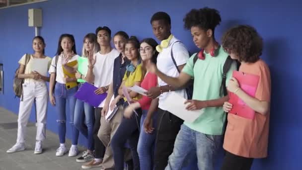 Grupo de amigos estudiantes adolescentes multiétnicos saludando diciendo hola a la cámara de pie sobre la pared azul en la escuela secundaria. Diversidad en la educación y la sociedad — Vídeos de Stock