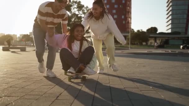Подростковые друзья веселятся вместе на скейтборде в современном городе на закате. Счастливая афроамериканская девочка сидит на коньках, которую толкает азиатский мальчик. — стоковое видео