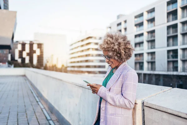 Hymyilevä Afrikkalainen Amerikan liiketoiminnan nainen afro hiukset käyttäen älypuhelin seisoo katolla auringonlaskun aikaan — kuvapankkivalokuva