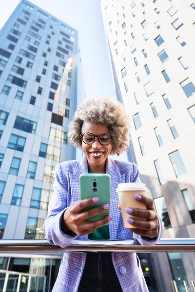 Munter ung, afroamerikansk forretningskvinne som holder kaffekopp mens hun bruker mobiltelefon i et finansdistrikt om morgenen – stockfoto