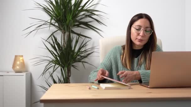 Młoda przedsiębiorczyni z okularami pracująca na laptopie i notatkami na notatniku w przytulnym biurze — Wideo stockowe