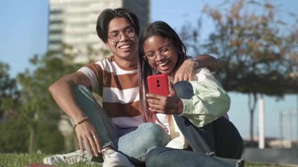Multiracial student par sitter på gräset på universitetsområdet gör online videosamtal med vän på smartphone — Stockvideo