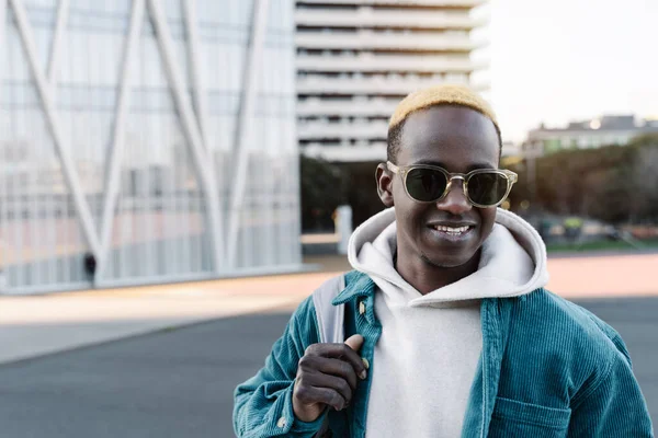 Smilende ung afrikansk mann med blondt hår og solbriller som går med pose i moderne by – stockfoto
