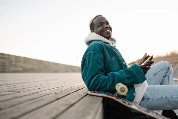 Kul, ung, afroamerikansk mann med skateboard hvilende ved siden av seg. – stockfoto