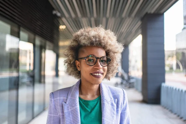 Smilende afroamerikansk forretningskvinne med afrohår som står i gangen utenfor bygningen om dagen – stockfoto