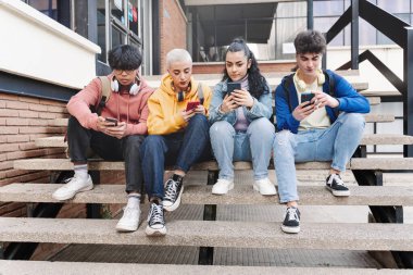 Çok kültürlü bir grup genç arkadaş cep telefonu ve sosyal medya kullanıyor. Üniversite kampüsünde oturan öğrenciler akıllı telefonlarda yazı yazıyor.