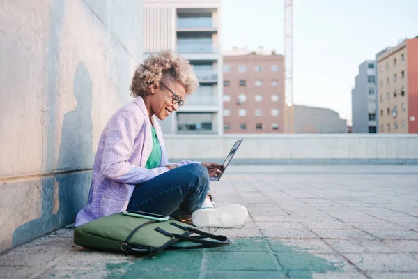 Lykkelig svart kvinne som jobber med laptop på bakken i byen utendørs. Moderne afroamerikansk forretningskvinne med blondt hår – stockfoto