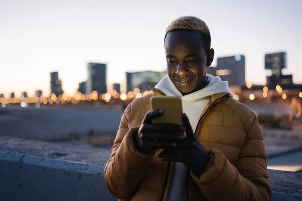 Afrikkalainen amerikkalainen mies käyttää älypuhelinta kaupungissa yöllä. Hymyilevä Cool musta kaveri matkapuhelimella sosiaalisen median, Verkkokaupoissa, Tekstaaminen Dating App — kuvapankkivalokuva