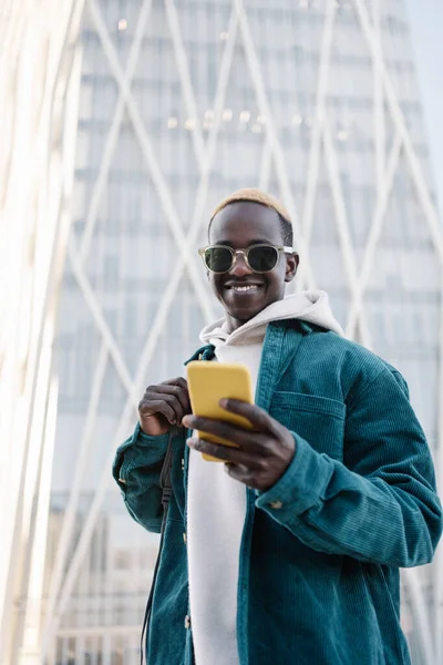 Smilende ung afrikansk mann med blondt hår og solbriller med pose og gul mobiltelefon – stockfoto