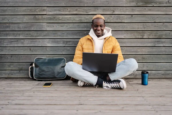 Afroamerikansk mannlig universitetsstudent som sitter utendørs og jobber med laptop og smiler med gul hettegenser – stockfoto