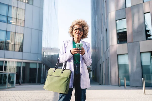 Smilende afroamerikansk forretningskvinne med smarttelefon stående med bag i byen på solskinnsdag – stockfoto