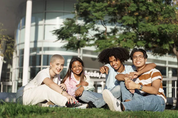 Unge, mangfoldige, glade studentvenner som sitter på gresset i byparken og ser mot kameraet – stockfoto